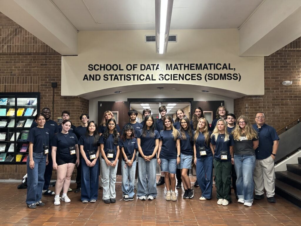 A group of students and two adults stand in front of a sign reading “School of Data, Mathematical, and Statistical Sciences (SDMSS)” inside a building.