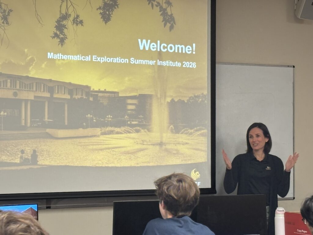 A woman stands in front of a projected presentation slide that reads “Mathematical Exploration Summer Institute 2026” while speaking to an audience.