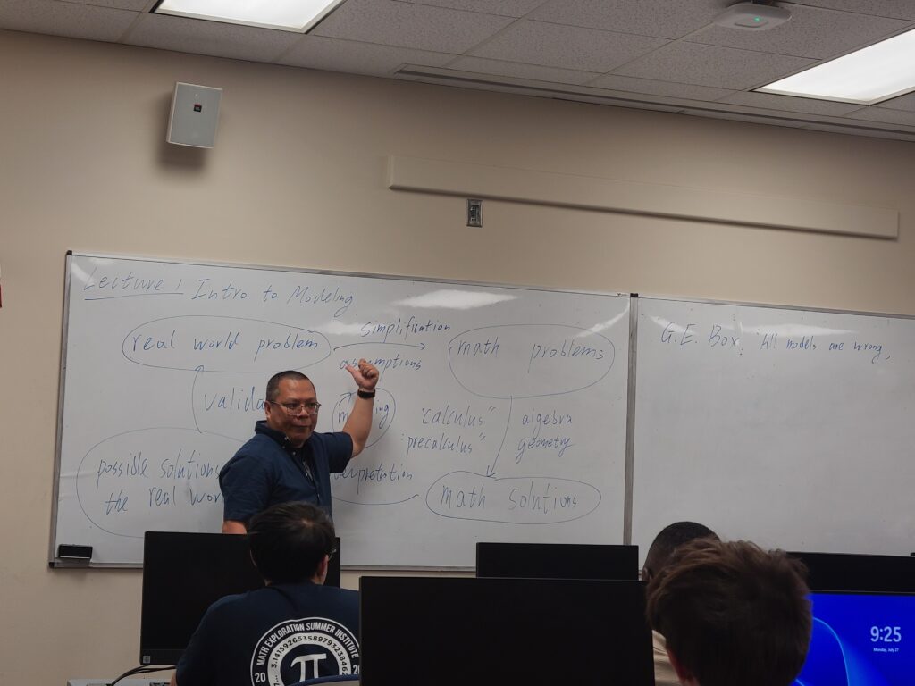 A teacher stands in front of a whiteboard with math terms written on it, addressing students seated at computer desks in a classroom.