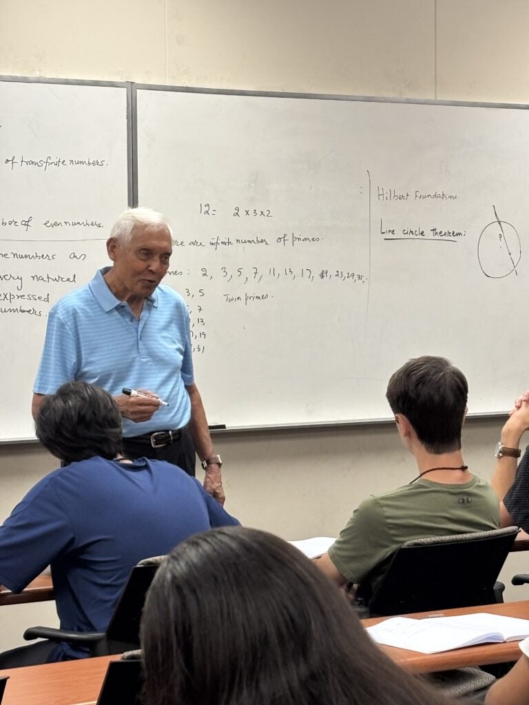 An older man stands in front of a whiteboard with mathematical notes, teaching a classroom of students who are seated and taking notes.