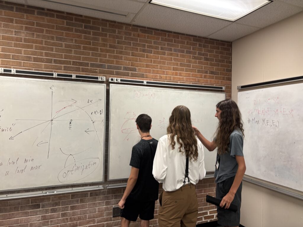 Three people stand and write on whiteboards filled with mathematical graphs and equations in a classroom with brick walls and a drop ceiling.