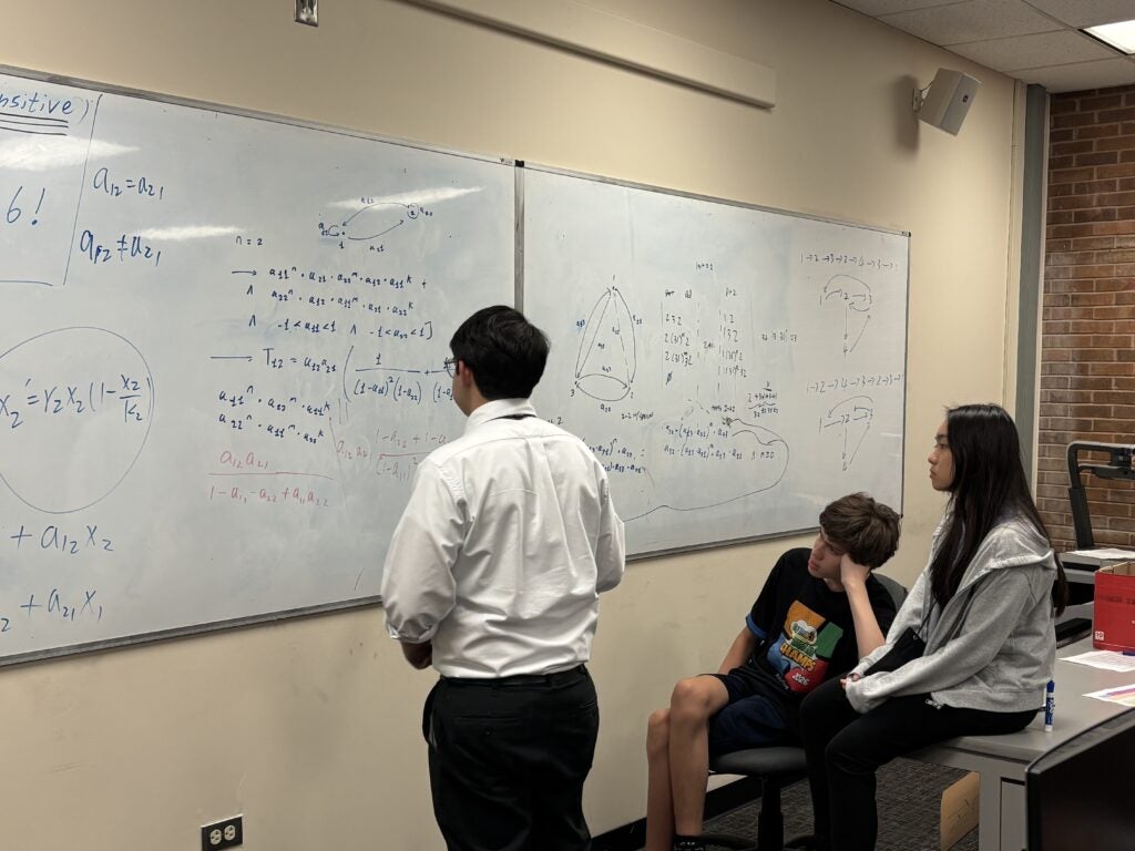 Three students focus on a whiteboard covered with complex mathematical equations and diagrams in a classroom setting.