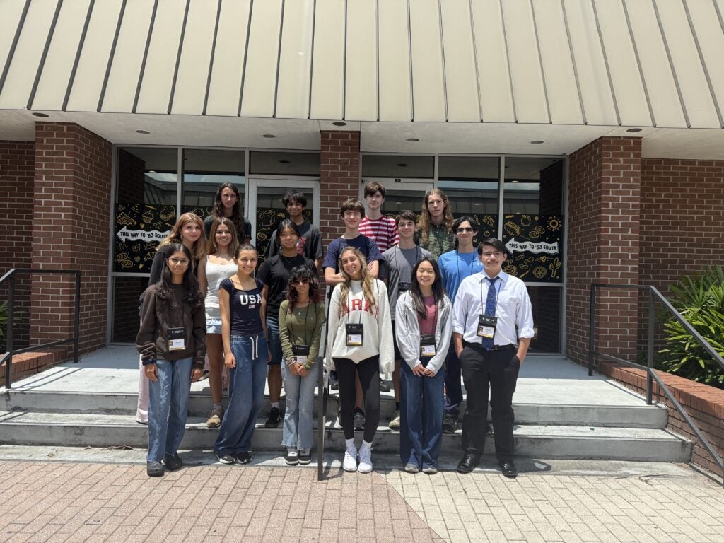 Sixteen teenagers pose on steps outside a brick building with glass doors, dressed in casual and semiformal attire, some wearing lanyards.