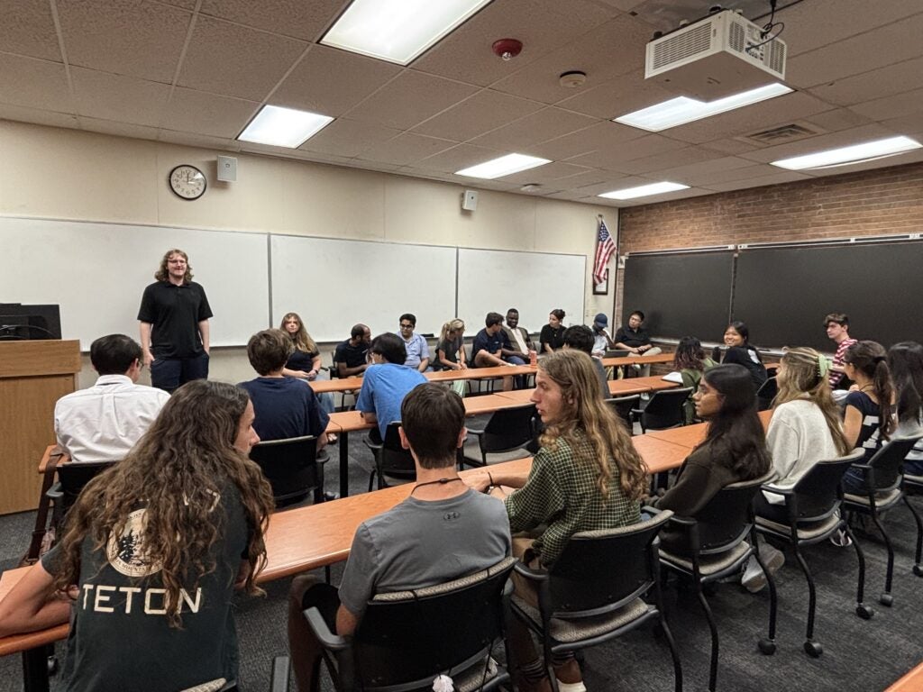A group of students sits around tables in a classroom, listening to a standing instructor near the front, with a U.S. flag and whiteboards in the background.