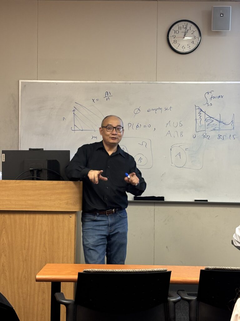 A person stands at a podium in a classroom, gesturing with a marker. Math equations and graphs are drawn on the whiteboard behind them. A clock on the wall shows 11:10.
