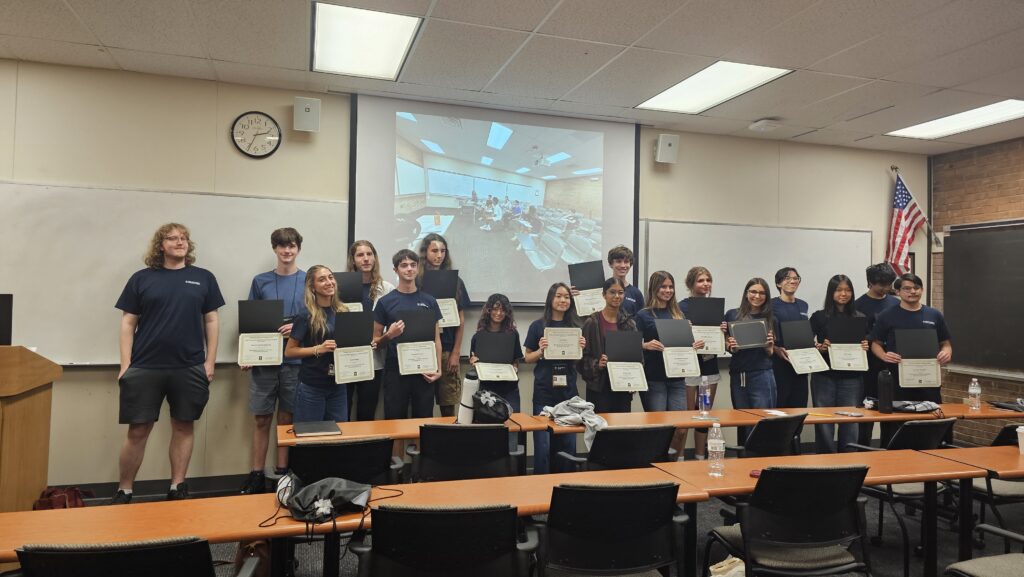 A group of students stands in a classroom holding certificates, posing for a photo in front of a projector screen and an American flag.