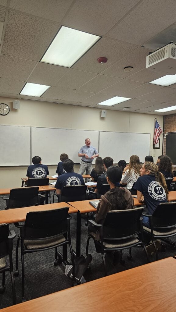 A teacher stands at the front of a classroom speaking to students, some wearing navy shirts with a white Pi symbol on the back. An American flag and whiteboards are visible.