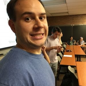 Man in a blue shirt making a nervous expression in a classroom with other people, some standing and some seated. One person is using a phone, and there is a chalkboard and a projector screen in the background.