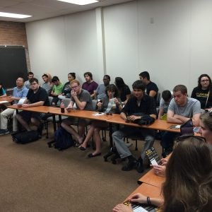 A group of students sits at long, connected desks in a classroom. Some are looking at papers or writing, while others are engaged in conversation. The room has white walls and a beige floor.