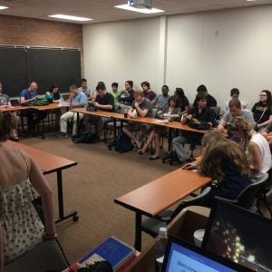 A group of students is seated in a classroom arranged in a U-shape. Many are using laptops and notebooks, with a projector screen slightly visible in the foreground.