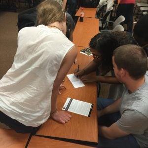 People are gathered around a table, with one person writing on a card while others observe.