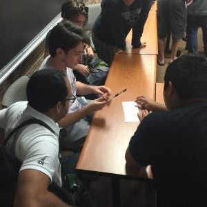 A group of students gather around a desk in a classroom, discussing and writing on a piece of paper. Some students are seated while others stand around them.