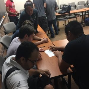 A group of people gathers around a table in a classroom, intently looking at a piece of paper and discussing its content.