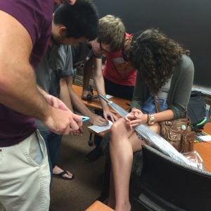 Group of people in a classroom closely examining a long object on a desk, with one person holding a smartphone.