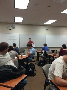 Students seated at desks in a classroom facing a whiteboard, where an instructor is speaking. Some students are taking notes while others listen attentively.