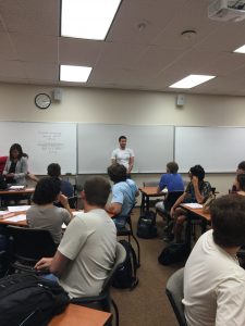 A man stands at the front of a classroom speaking to a group of seated students. A whiteboard with notes is visible behind him. Other people are seated and appear to be listening or working.