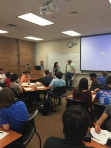 Classroom scene with students seated at tables, facing a whiteboard. Three individuals are standing at the front of the room, one of them speaking. A projector screen displays a blue background.