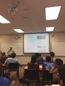 A classroom with a lecturer presenting a slide on the screen to seated students. The slide appears to contain text and images. The room has whiteboards, a clock, and ceiling lights.