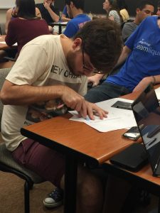 A person in a t-shirt and shorts is sitting at a desk, leaning over papers and writing. They have a laptop and a phone on the desk. Other people are seated in the background.