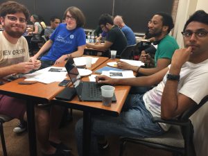 A group of people sitting around a table with laptops, papers, and food, engaged in discussion at a classroom setting.