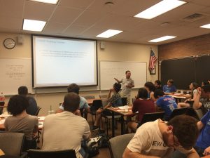 A classroom with students seated at tables, listening to an instructor who is standing next to a screen displaying a presentation. An American flag and a whiteboard with notes are in the background.