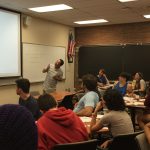 A teacher is standing in front of a classroom, pointing at a projection screen. Students are seated at desks, attentively looking towards the teacher and the screen. An American flag hangs on the wall.