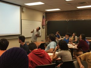 A teacher is standing in front of a classroom, pointing at a projection screen. Students are seated at desks, attentively looking towards the teacher and the screen. An American flag hangs on the wall.