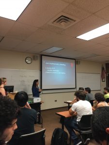 A woman is presenting a slide titled "Joint Mathematics Meeting" to a classroom of students. The slide includes details about a meeting scheduled for January 5th in Seattle, Washington.