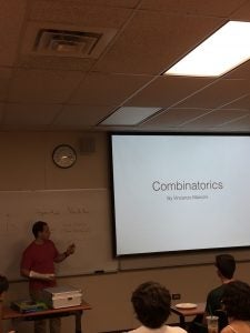 A man stands in front of a classroom holding a pointer, next to a projector screen displaying "Combinatorics By Vincenzo Marconi". Students are seated and listening.