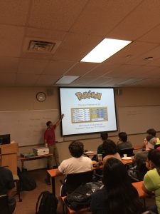 A person giving a presentation on Pokémon in a classroom, with a slide displaying "Choose a Pokémon out of 151" and listing the total number of Pokémon. Attendees are seated and looking at the screen.