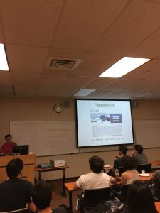 A person giving a presentation in a classroom. The slide on the screen is titled "Passwords" and includes a table and diagram. Several audience members are seated and watching.