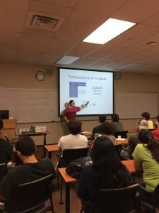 A man in a red and white shirt is giving a lecture on "Permutations of multiset" to a classroom of students, pointing to a slide with mathematical content.