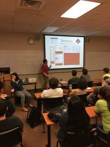 A person stands in front of a classroom, presenting a slide labeled "Dominos" with various menu options. Students are seated at desks, viewing the presentation.