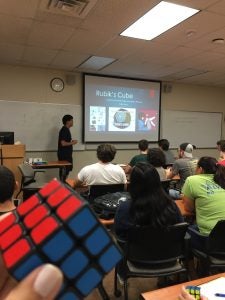 A person holds a solved Rubik's Cube in the foreground of a classroom where a presentation on the Rubik's Cube is being given to students.