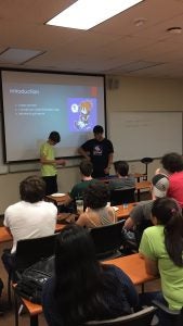 Two individuals present information about a Rubik's cube to a seated audience in a classroom. A projection screen displays an introduction slide.