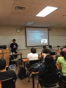 A person stands at the front of a classroom giving a presentation. A slide titled "Number of combinations" is displayed on the screen, and students are seated at desks, listening attentively.