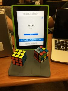 A tablet on a stand displays "Space Available" for UCF CMS Math. The screen shows a time and date. Two Rubik's cubes are placed in front of it. A laptop and another device are partially visible.