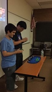 Two individuals stand at a table solving Rubik's Cubes in a dimly lit room. An American flag and a whiteboard are visible in the background.