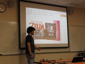 A person giving a presentation in a classroom. The screen displays the title "Western Music Today: The Legend of Zelda" with related images. The room has a clock and a whiteboard.