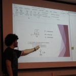 A person with dark curly hair is pointing at a projection screen displaying mathematical equations and variables related to string tension, mass, and length in a classroom setting.