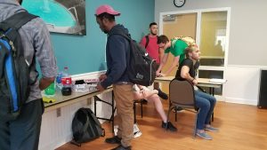 Several people are gathered in a room. One person is taking a slice of pizza from a table with pizza boxes and soda. Others are sitting or standing nearby, some with backpacks.