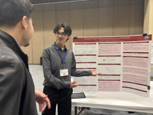 A person presents a scientific research poster to another attendee at an indoor conference or academic event.