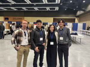 Four young adults wearing name badges and business casual attire stand together and smile in a large convention hall with booths and other people in the background.