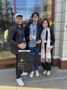 Three people stand outside a building holding drinks and wearing name badges. One holds a black shopping bag. They are dressed in casual winter clothing.