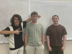 Three young men stand in front of a whiteboard. The person on the left points at the center person, who is being patted on the head. The person on the right stands with hands at sides.