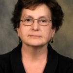 A woman with short, curly brown hair, glasses, and a neutral expression is wearing a dark top in front of a dark, blurred background.