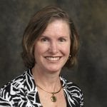 A woman with shoulder-length brown hair smiles for a portrait against a neutral background. She is wearing a black-and-white patterned outfit with a gold necklace.