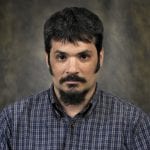 A man with a serious expression, dark hair, a beard, and a plaid shirt poses against a neutral, slightly textured background.