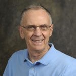 An older man with glasses and short gray hair, smiling, wearing a light blue collared shirt, against a dark mottled background.