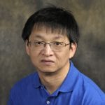 A headshot of a person wearing glasses and a blue polo shirt, set against a dark, textured background.